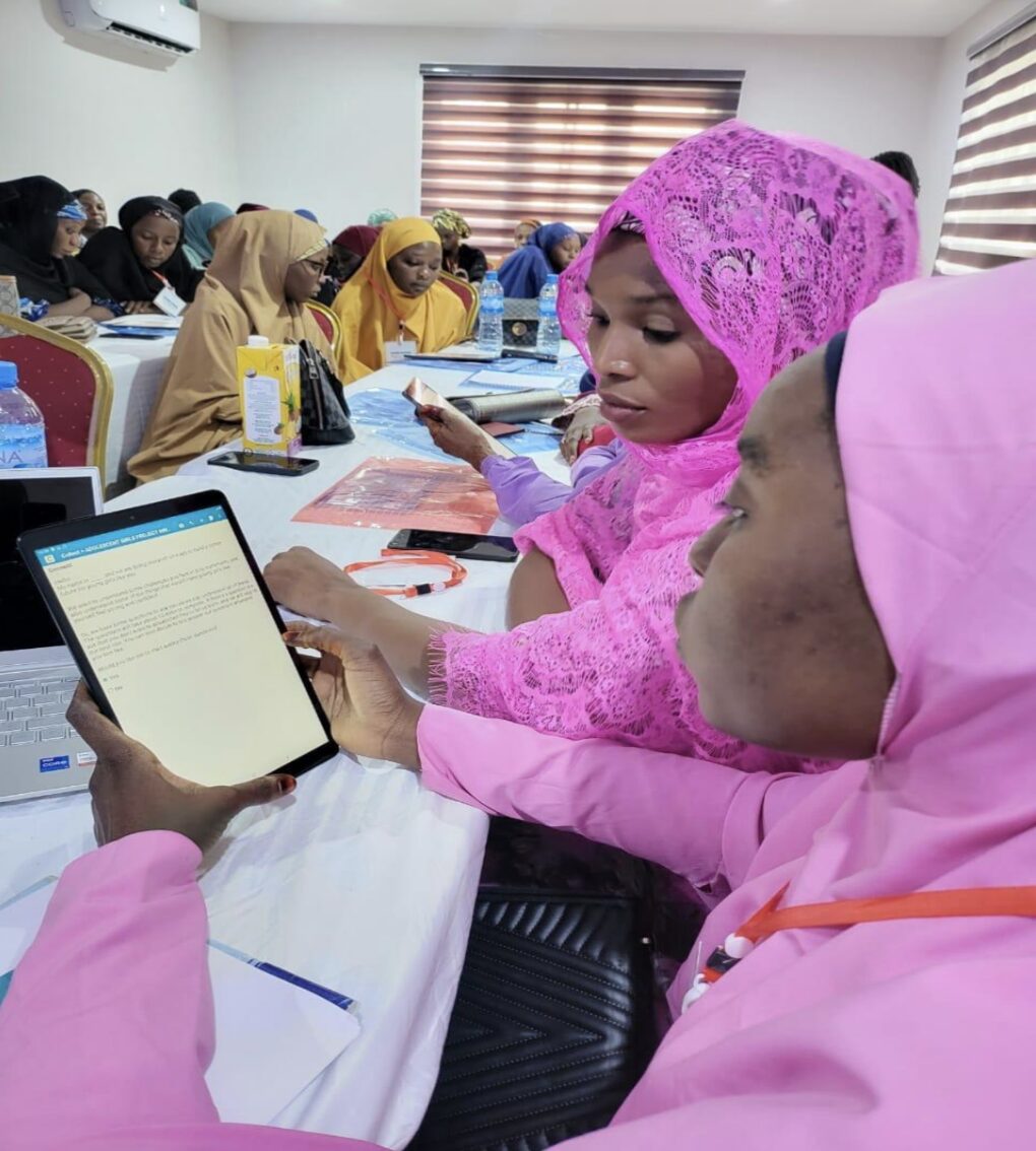 A group of the young women researchers sitting at long tables, working on documents and devices. They are receiving training on ethnographic methodologies and research skills. 