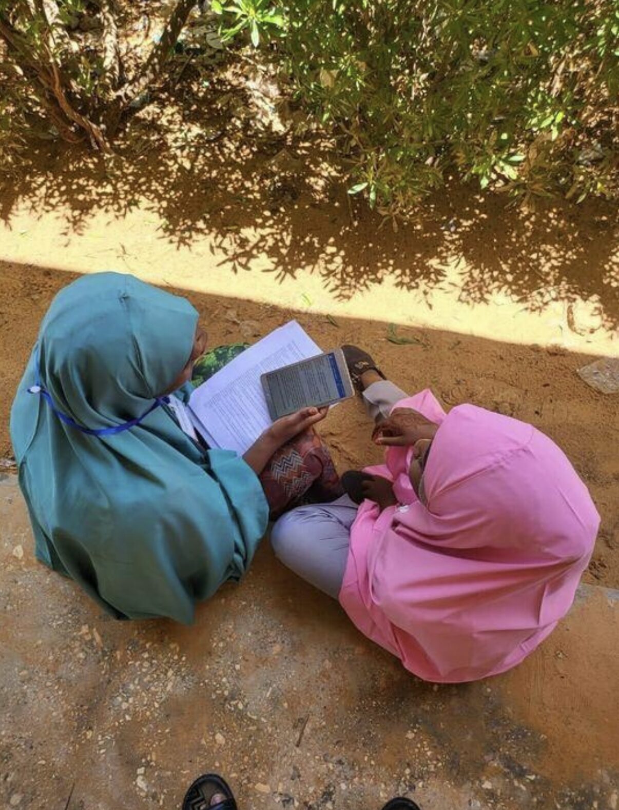 A young woman researcher sitting beside a research participant (an adolescent  girl). The researcher is londing documents and a tablet device, and is administering a quantitative survey.