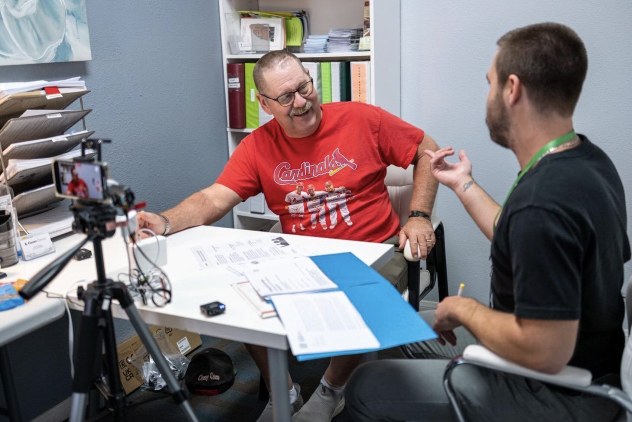 A researcher and participant sit at a desk having a conversation. The smiling participant is an older adult with glasses, a mustache, and a Saint Louis Cardinals t-shirt. The researcher wears jeans and a black t-shirt, and sits with back to the camera. A folder is open on the table between the two, and a small camera on a stand records their conversation.