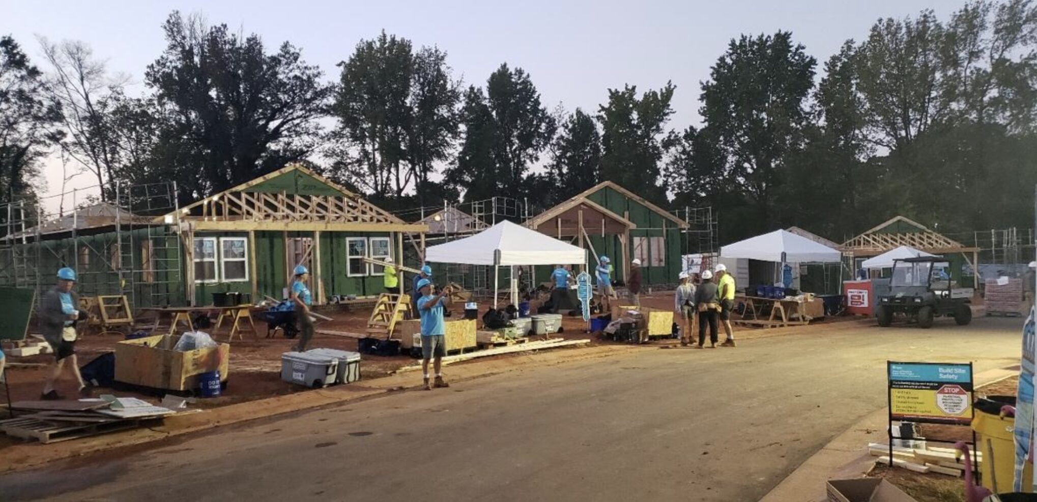 Street view of homes under construction in a cul-de-sac. Visible outside the homes are volunteers in hard hats, construction materials, and white canopies.