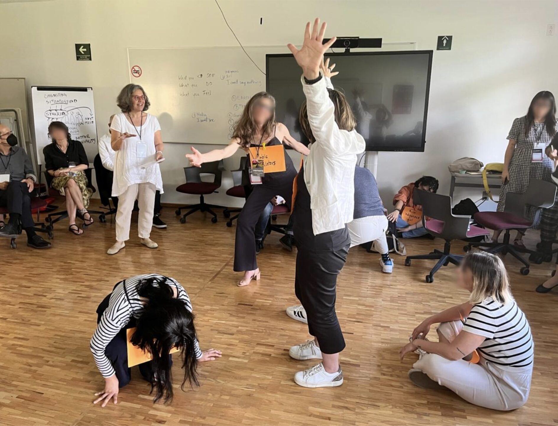 An interior room view with a group of participants creating a circular human sculpture, the participant in the center is in standing position with her arms raised and palms opened, on the left, another participant is in a squatting crouched position, while on the right, one, is calmly watching the scene in a confortable sitting position. On the back another participant is lightly inclined facing towards the center with his arms open while in the far left, the facilitator is standing watching the scene.