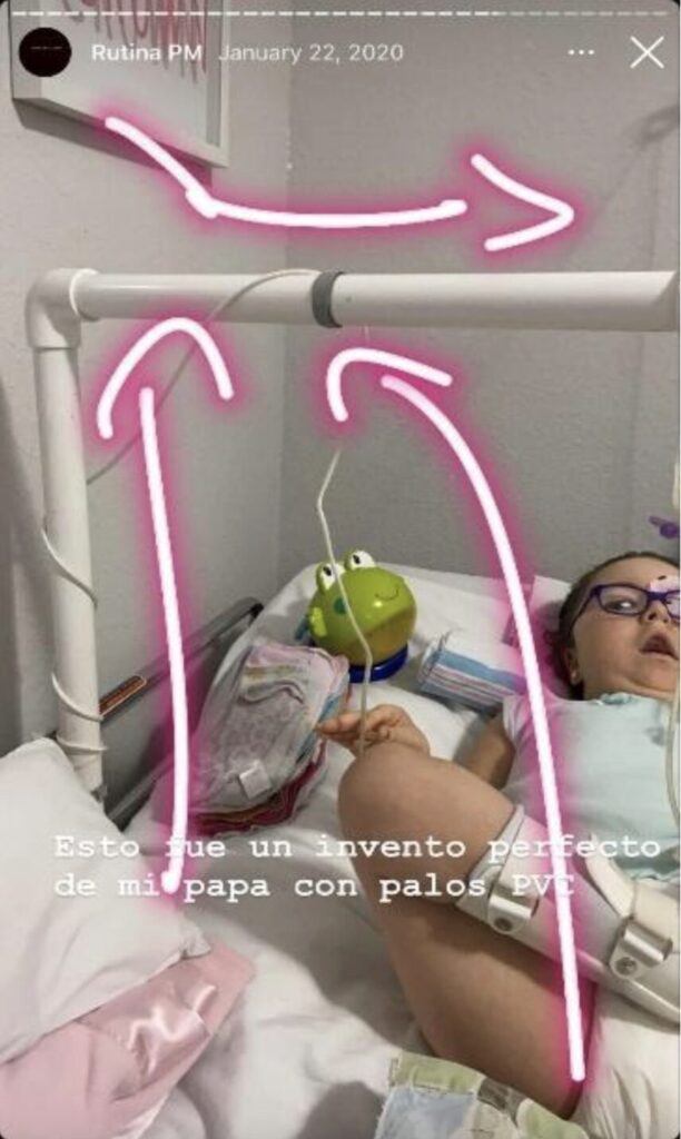 Instangram post shows a photo of a child with SMA in bed with a toy and blankets. Bright pink arrows indicating the PVC pipe structure with cables wound around it. and the following text, are overlain on the photo: “Esto que un invento perfecto de mi papa con palos PVC.” At the top of the pos tis the text “Runia PM, January 22, 2020.”
