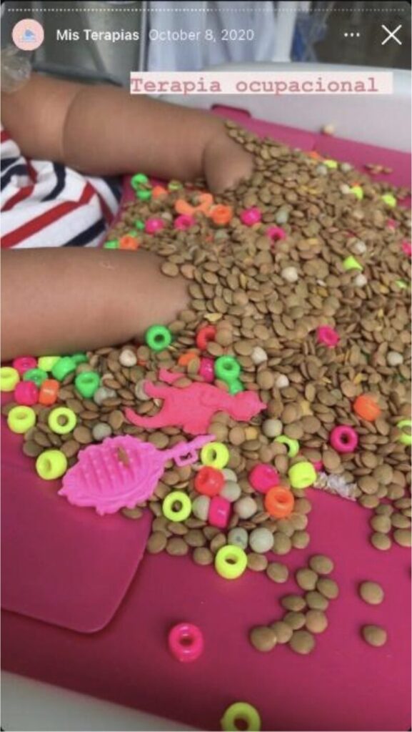 A close-up photo of a child’s lap, covered with a pile of dried lentils, beads, and small plastic figures. The child’s hands are nestled within the pile.